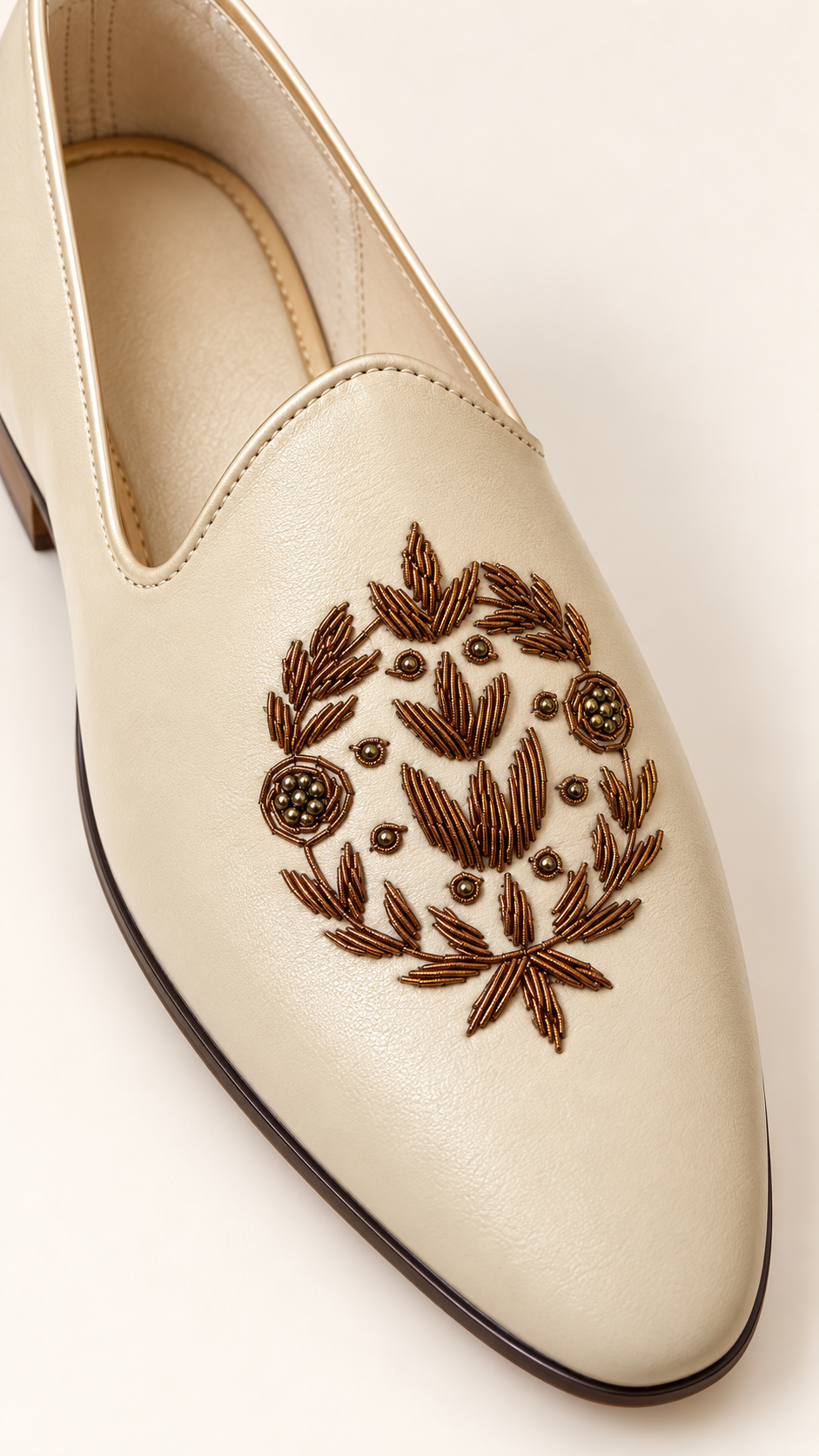 A close-up of a cream-colored leather slip-on shoe featuring an intricate brown embroidered floral wreath design on the toe box, set against a solid off-white background.