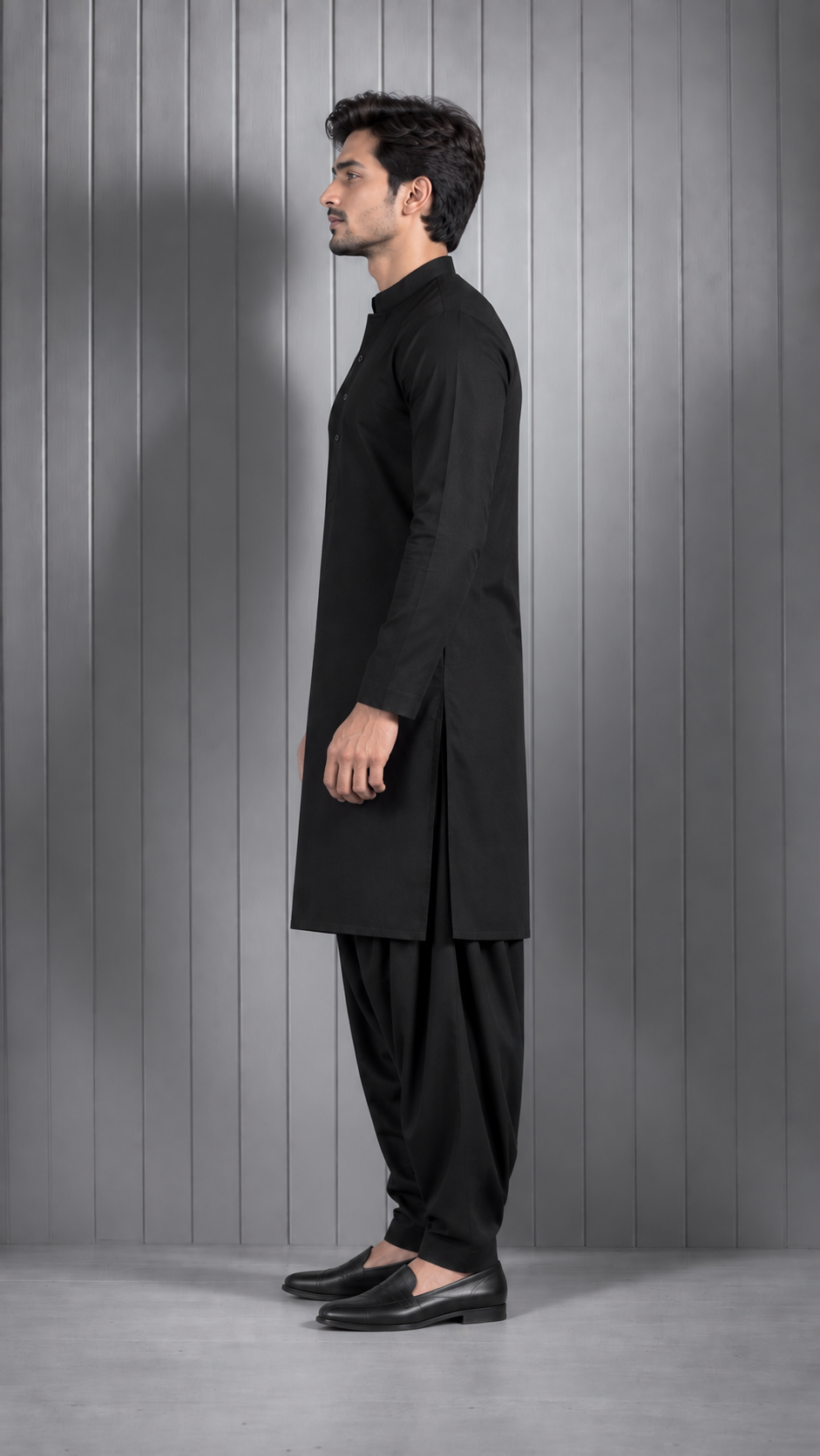 A man in a black long kurta and black pants stands in profile against a gray paneled studio wall.
