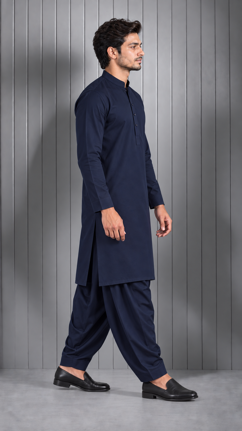 A man wearing a dark navy blue traditional kurta and pant set with black loafers, standing in profile against a gray paneled studio wall.
