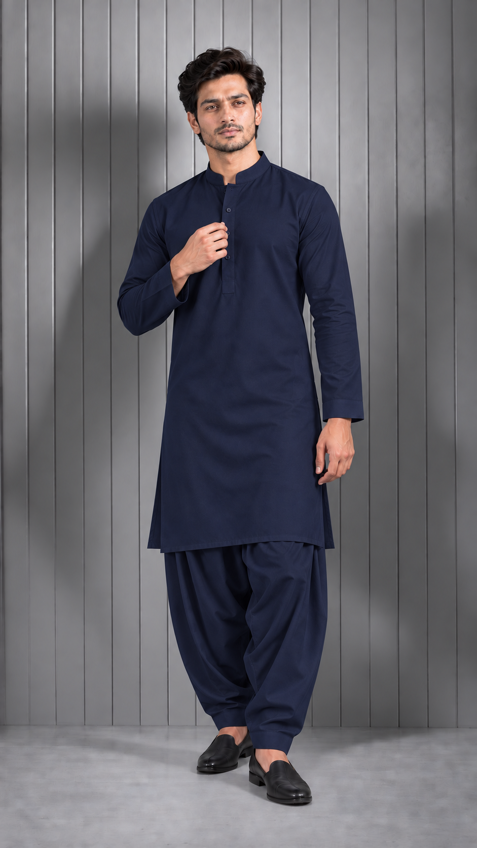 A man modeling a dark navy blue traditional Indian outfit consisting of a long-sleeve kurta and matching pants, standing against a gray paneled studio background.