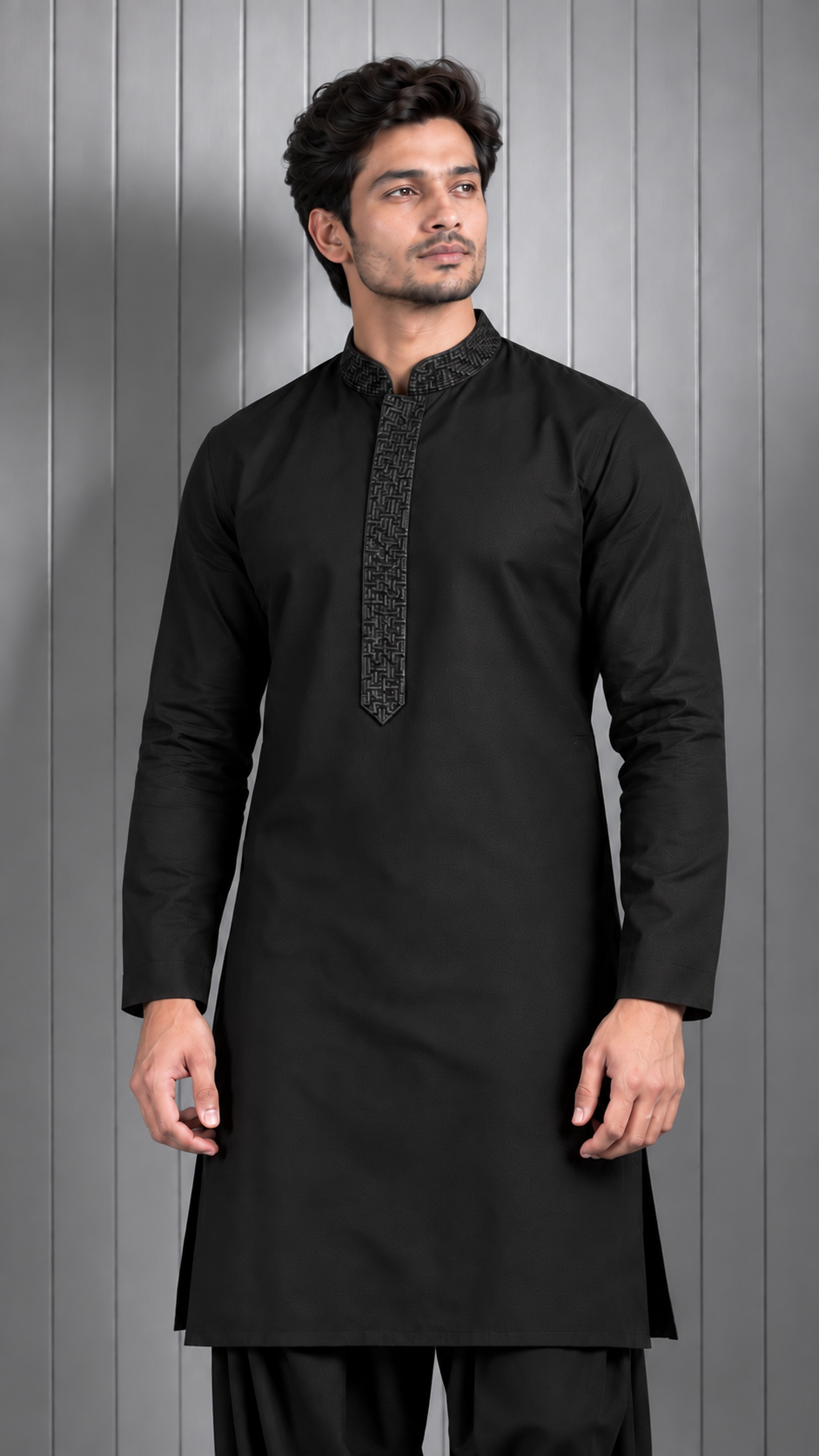A man wearing a black long-sleeve kurta with a textured patterned placket and collar, standing against a gray paneled background.