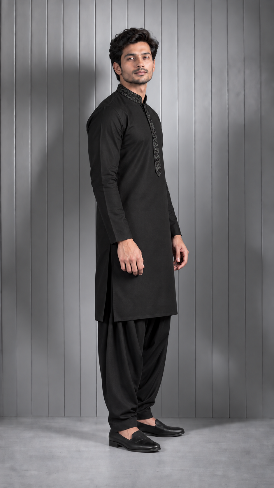 A man modeling a black long-sleeve kurta with matching black pants and black loafers, standing in profile against a gray paneled studio wall.