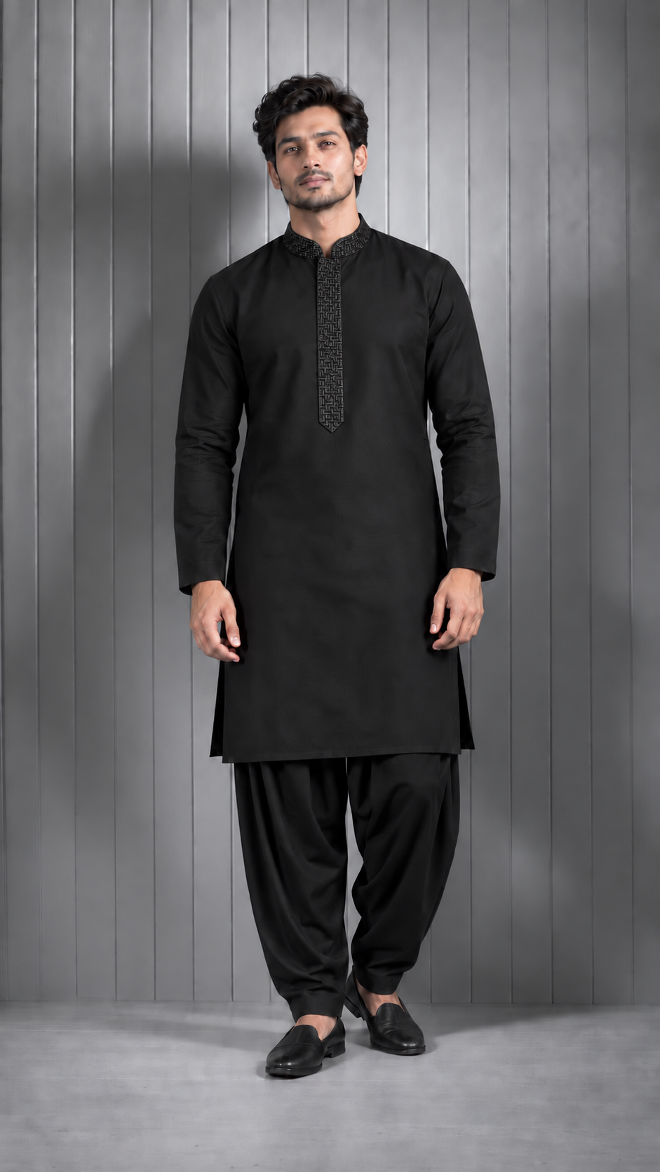 A man modeling a black long-sleeve kurta with a patterned placket and black shalwar pants, standing against a gray paneled studio background.
