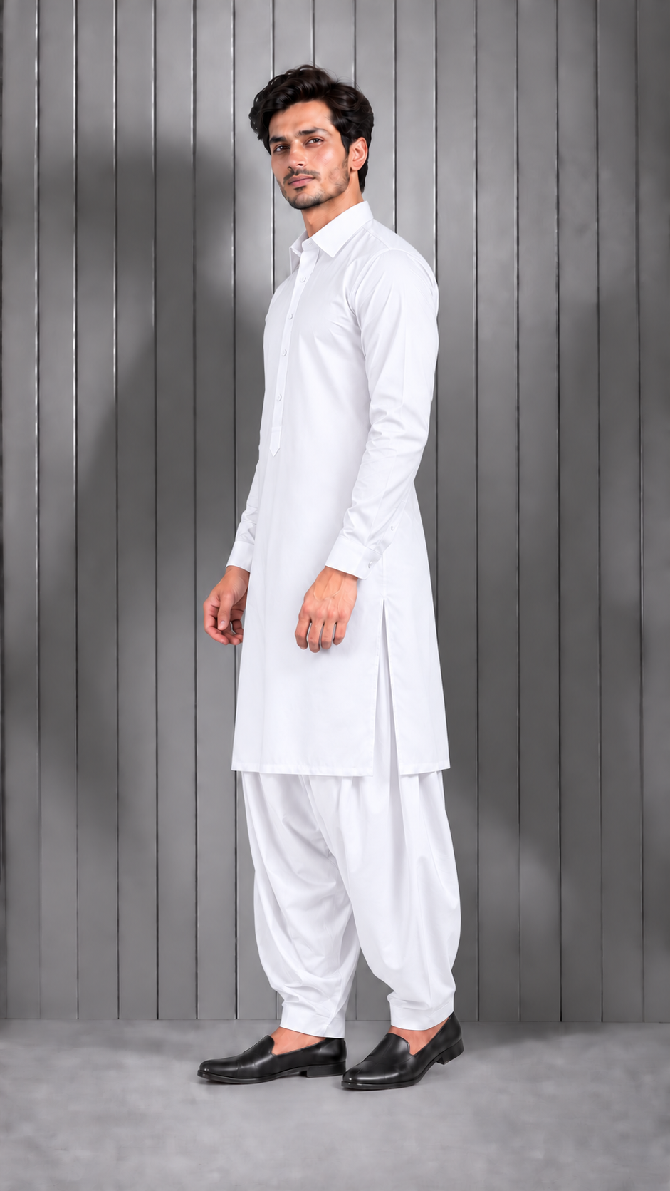 A man modeling a white long-sleeve kurta and white pant suit with black loafers, standing against a gray vertical wood-paneled wall.