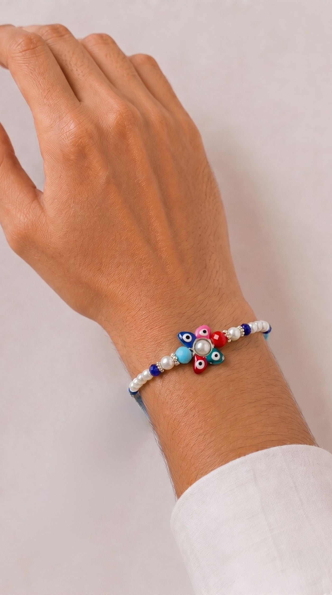 Multicolour evil eye flower rakhi bracelet with pearl centerpiece, colourful beads, white pearl accents, and adjustable navy blue thread worn on a man's wrist – Diya Onlinets