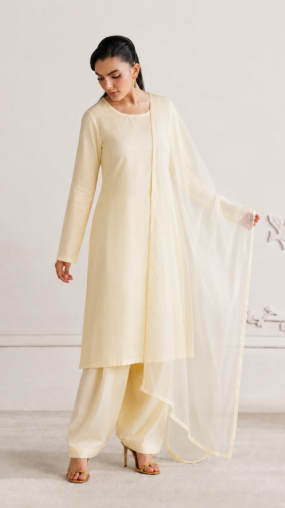 A woman modeling a cream-colored traditional outfit consisting of a long kurta, matching pants, and a sheer dupatta, standing against a light beige wall.