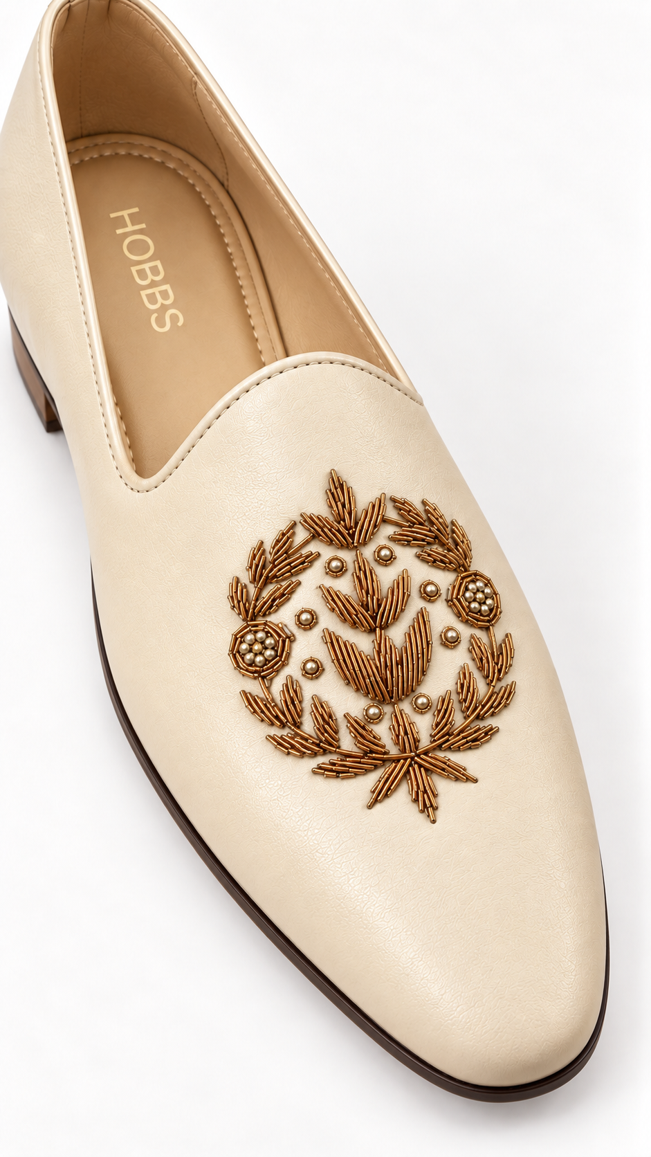 A cream-colored leather slip-on loafer with a brown embroidered laurel wreath design on the toe, featuring the brand name 'HOBBS' printed on the inner sole, set against a plain white background.